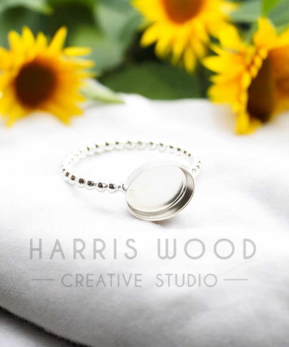 A silver ring with a smooth oval bezel, displayed on a white fabric with sunflowers in the background. The ring appears to be lightweight and solid silver.